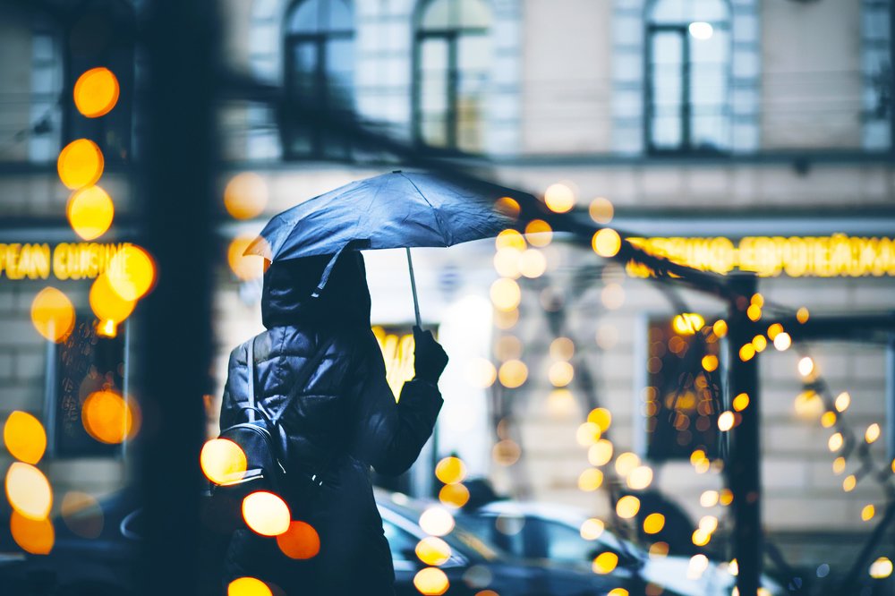 A woman in a black windbreaker with a wet umbrella walks along a city street in the evening