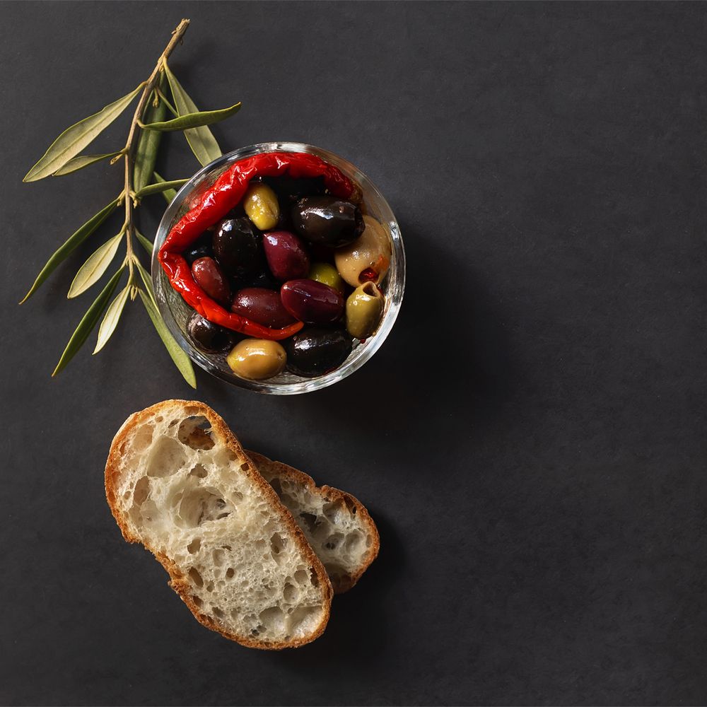 Bowl with different types of olives, ciabatta bread and olive twig on dark background.