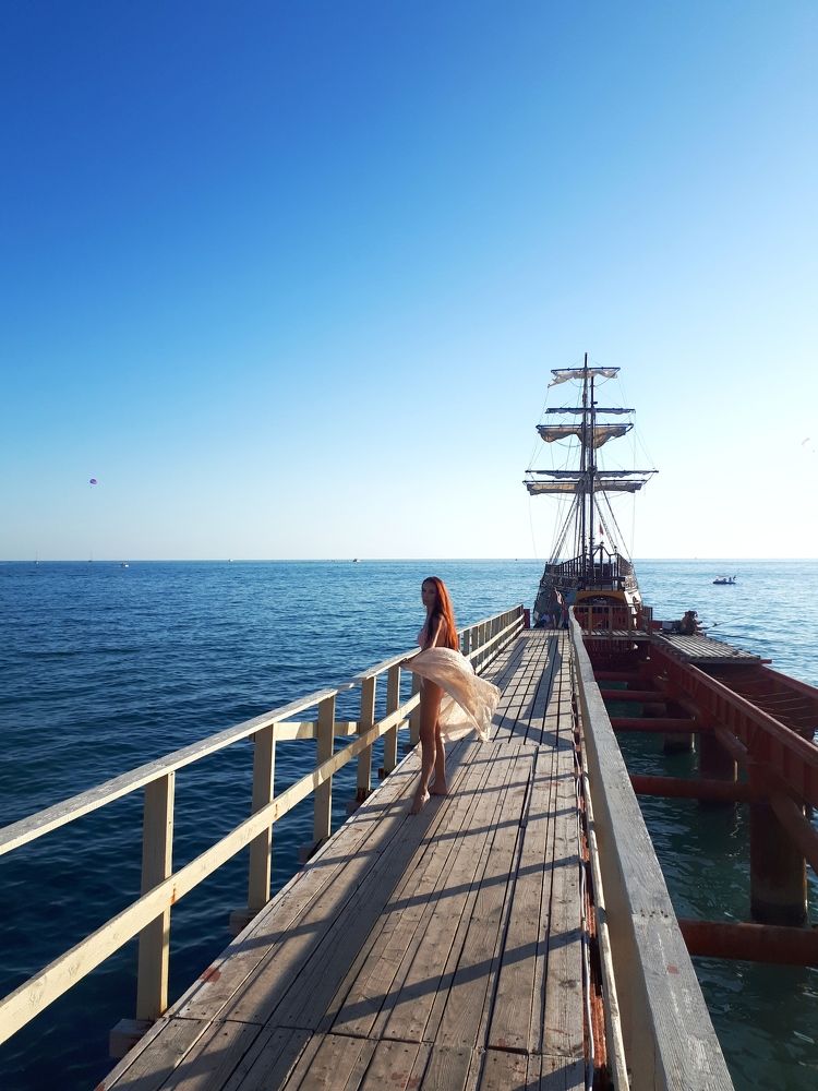 A girl stands on a small pier in the background of a sailing ship.
