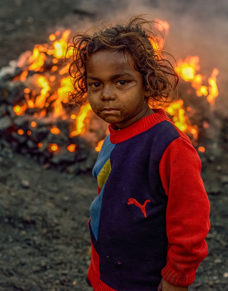 A child with coal smudged face standing in front of fire made for charcoal preparation