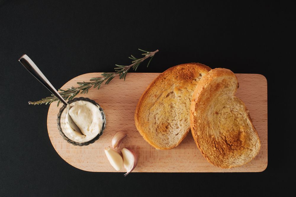 Wooden kitchen board with toasted bread, spread, onions and rosemary on black background.
