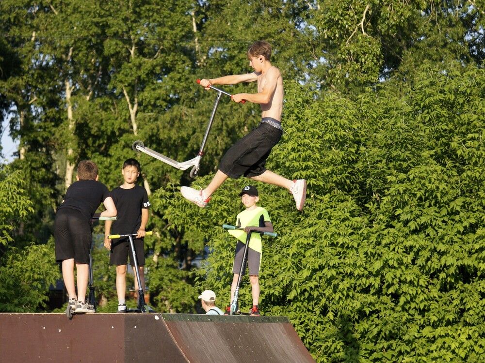 A teenager demonstrates high jump tricks on a scooter against the background of trees in a skatepark.