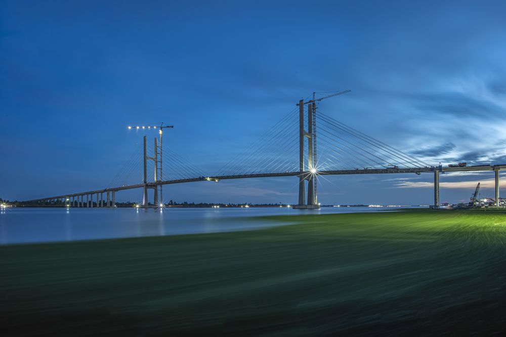 A ribbon of green silk drapes across the ongoing cable-stayed bridge construction site.