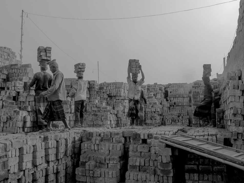 A brickfield worker pushes a handcart loaded with freshly molded clay past towering stacks of sun-dried bricks, as smoke rises from the kiln chimney in the background. In Bangladesh’s brick-making yards, laborers spend long hours shaping, carrying, and fi