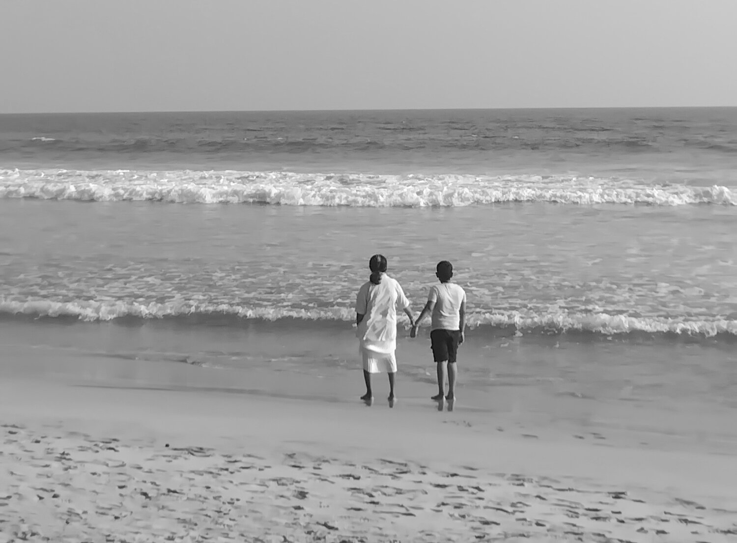 A serene, lighter-toned monochrome image of an adult and a child standing in the shallow surf.