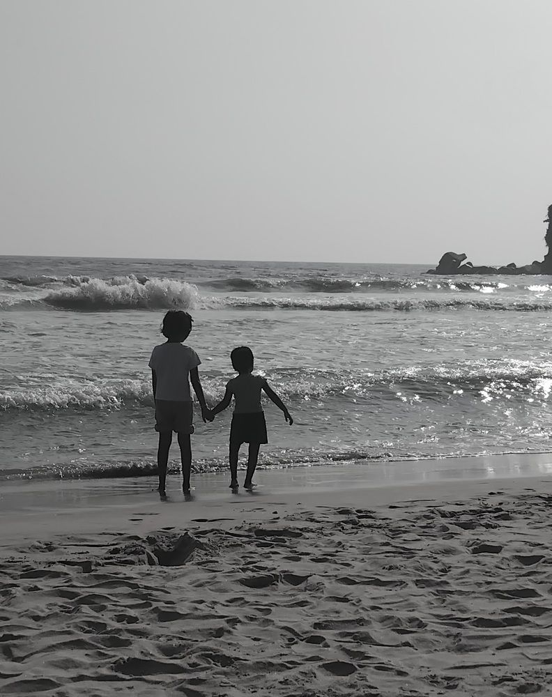 ​A high-contrast, black-and-white shot of two young children standing at the water\\\'s edge.