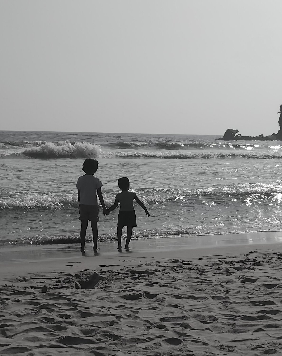 ​A high-contrast, black-and-white shot of two young children standing at the water\\\'s edge.