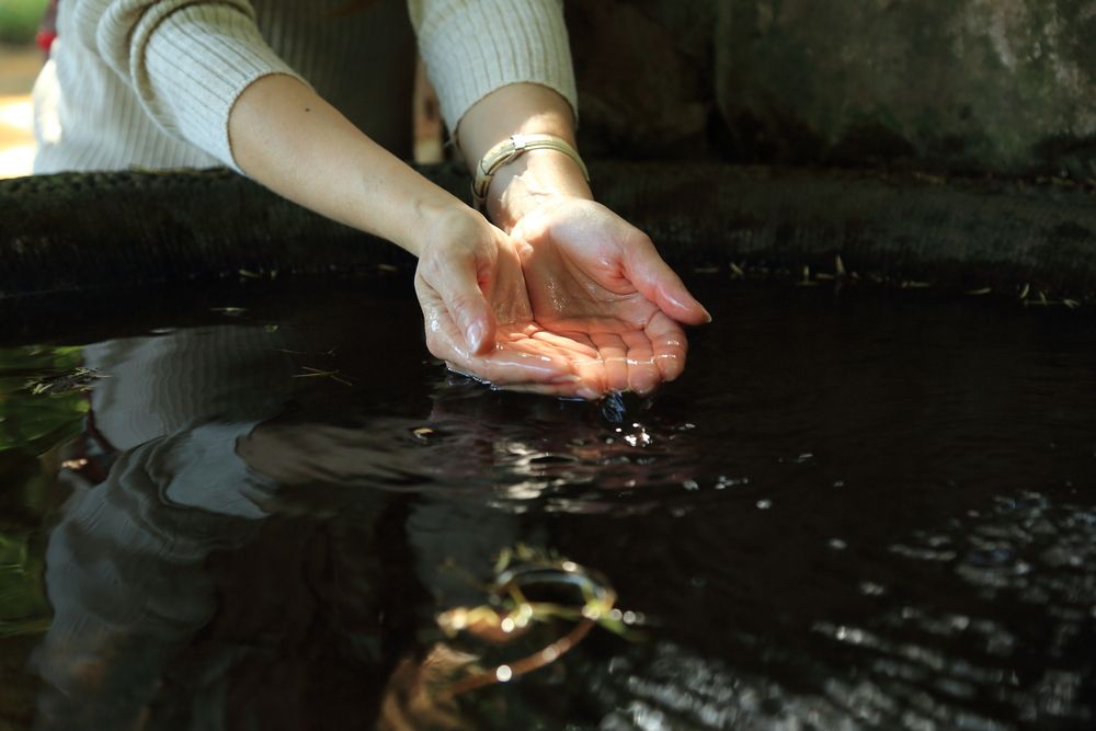 A close up shot of female hands cupping clean fresh water from a dark stone source fountain outdoors in summer