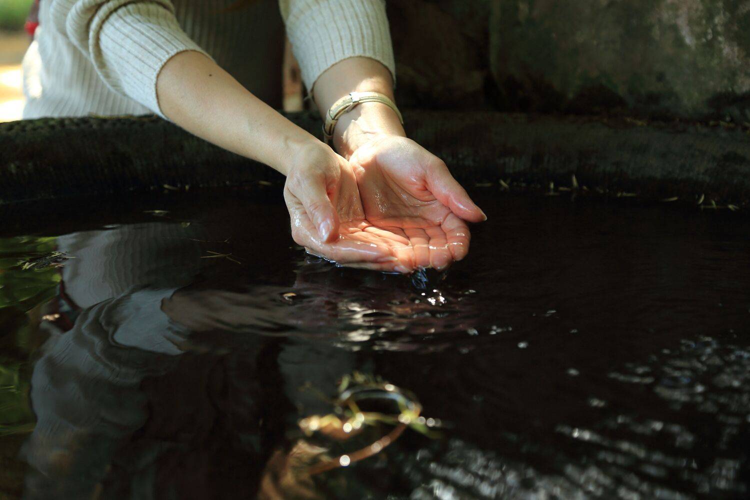 A close up shot of female hands cupping clean fresh water from a dark stone source fountain outdoors in summer