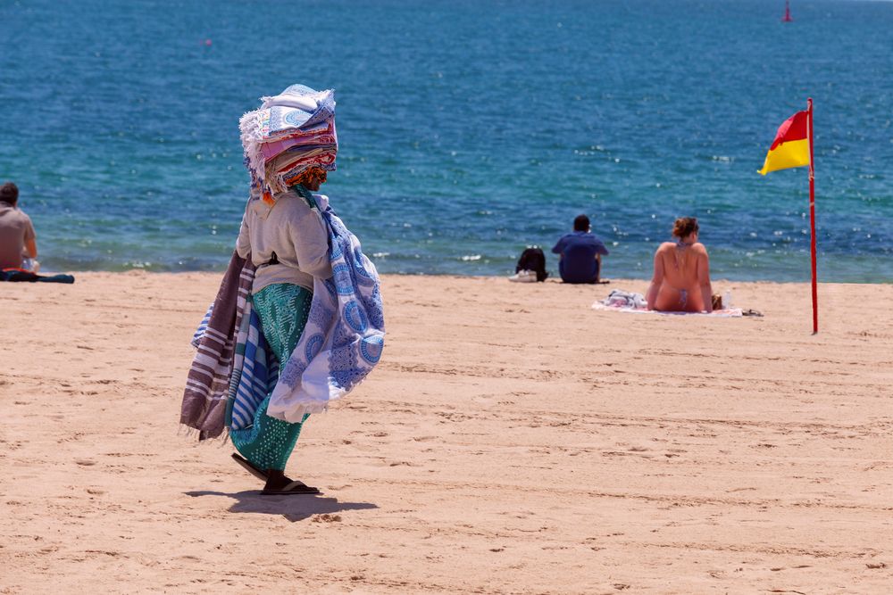 A beach vendor carrying colorful textiles walks along Cascais Beach