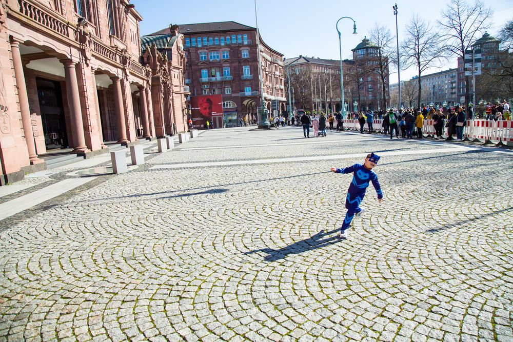 A spectacular wide angle photograph showing a young child in a blue carnival costume running on a massive stone pavement square near historic buildings