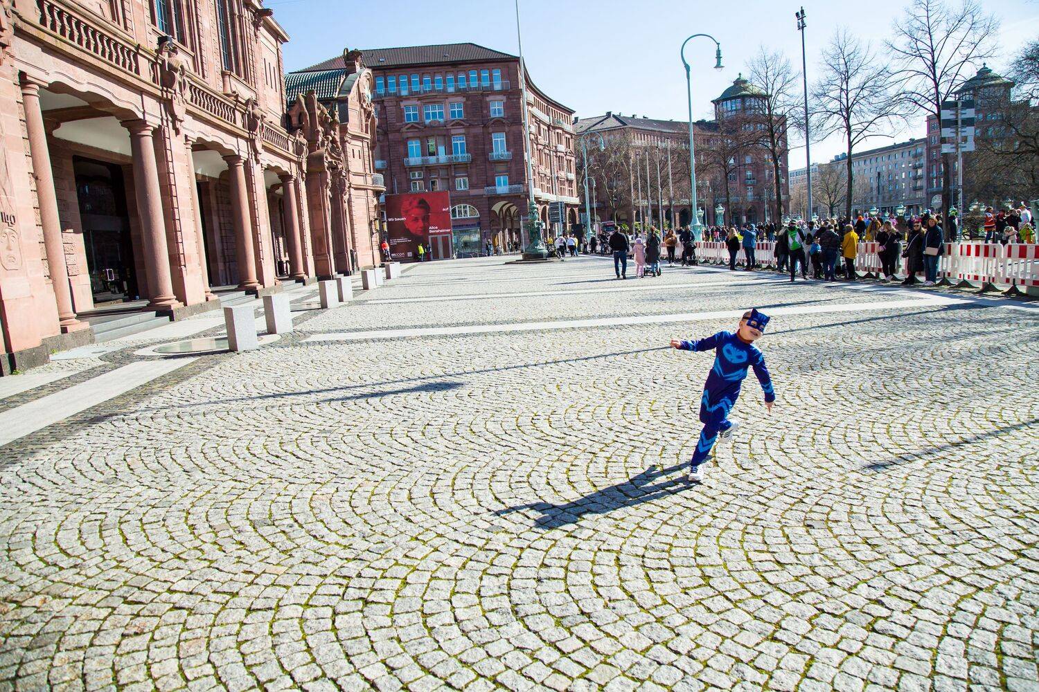 A spectacular wide angle photograph showing a young child in a blue carnival costume running on a massive stone pavement square near historic buildings
