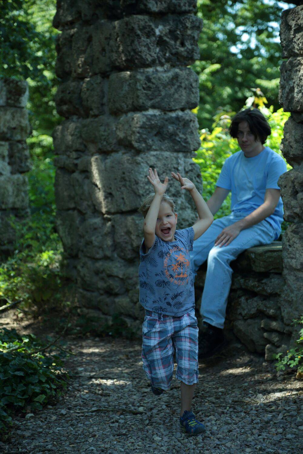 A beautiful vertical lifestyle photograph showing a joyful little boy running and cheering near ancient stone ruins while another young person sits in the background