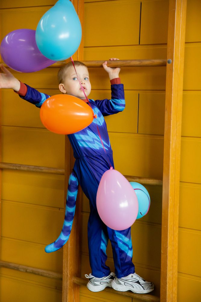 A vertical medium portrait photograph focusing on a young child wearing a blue superhero carnival costume holding colorful balloons next to wooden wall bars