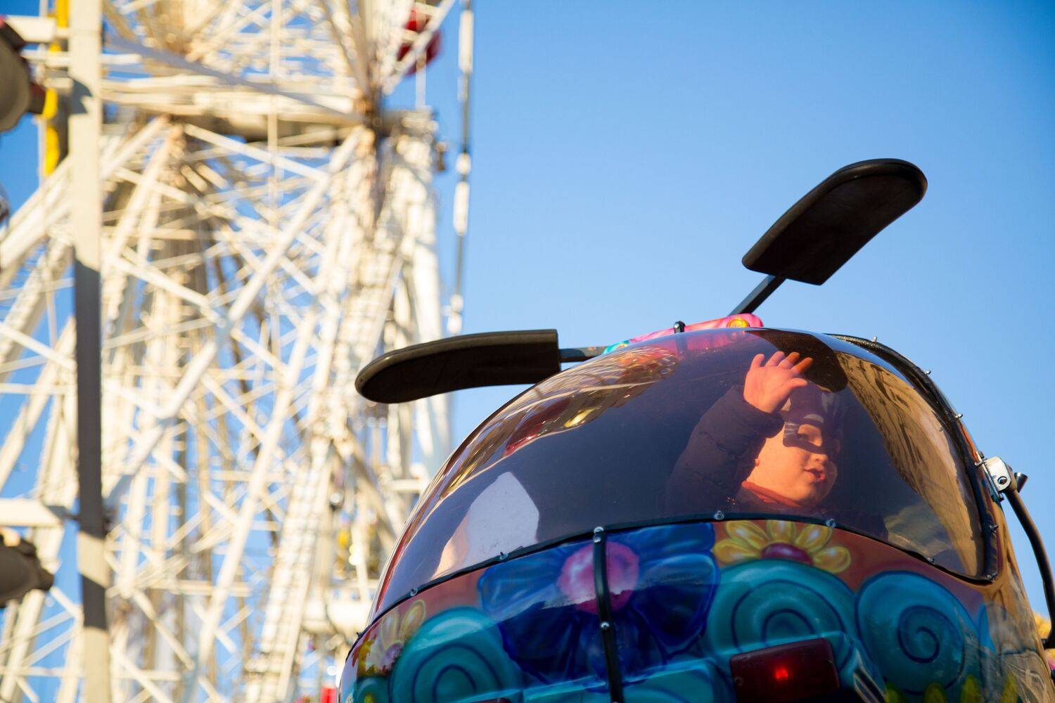 A beautiful low angle photograph focusing on a happy child waving from a colorful helicopter amusement ride cabin with a massive white ferris wheel background