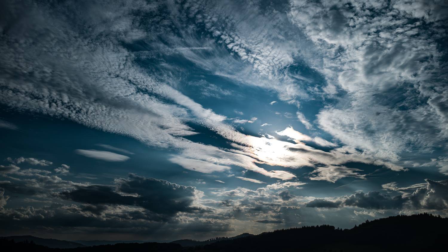A magnificent high angle aerial drone photograph focusing on a dramatic cloudy blue sky with bright sun rays breaking through texture layers over a dark picea abies spruce forest silhouette