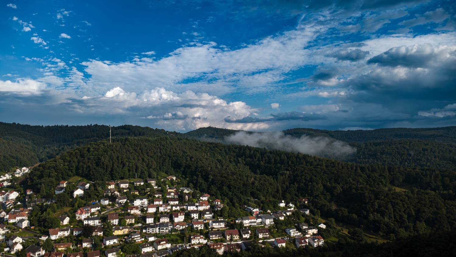 A magnificent high angle aerial drone photograph showing a scenic European village surrounded by dense picea abies spruce forests under a dramatic cloudy blue sky