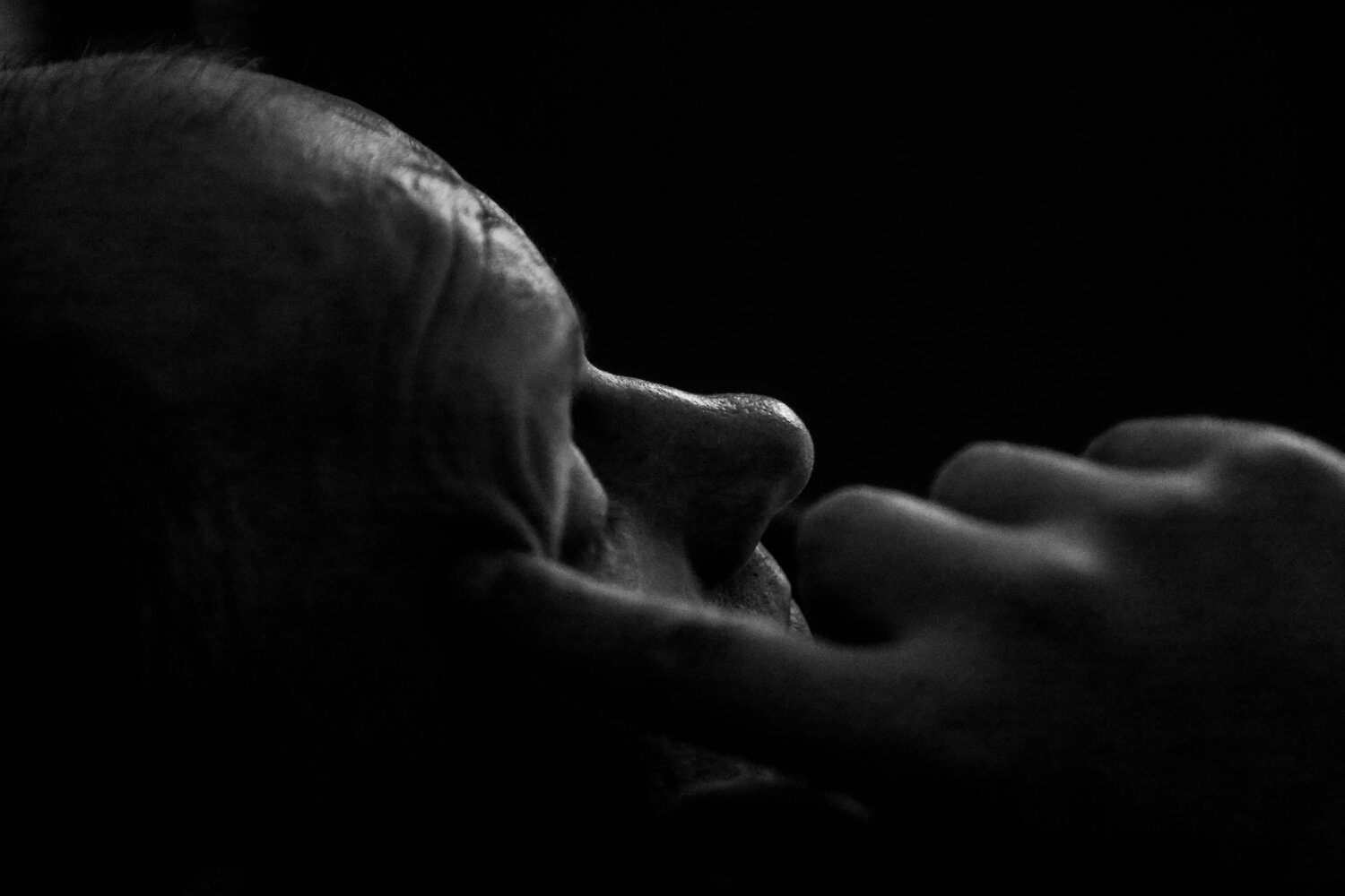 An emotional dramatic black and white portrait photograph showing a side profile view of a thoughtful senior adult man homo sapiens touching his face in deep contemplation