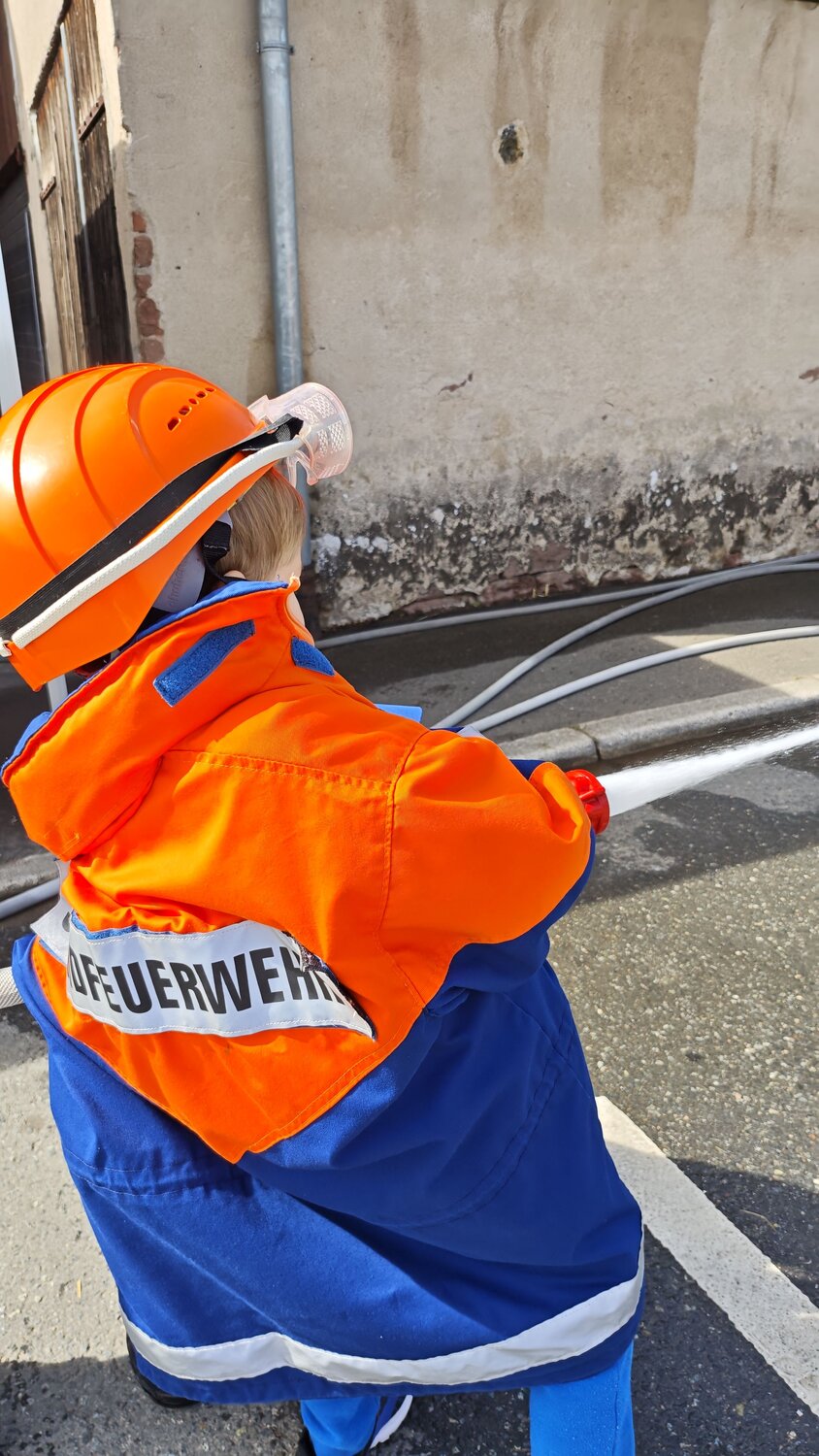 A detailed back angle photograph showing a young boy wearing a bright orange firefighter protective helmet and uniform while spraying water with a hose in a street near an old building wall