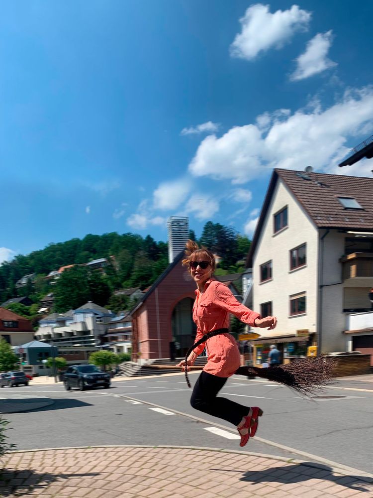 A cheerful young woman in sunglasses jumps high mimicking flying on a traditional wooden broomstick near green trees and houses under a blue sky
