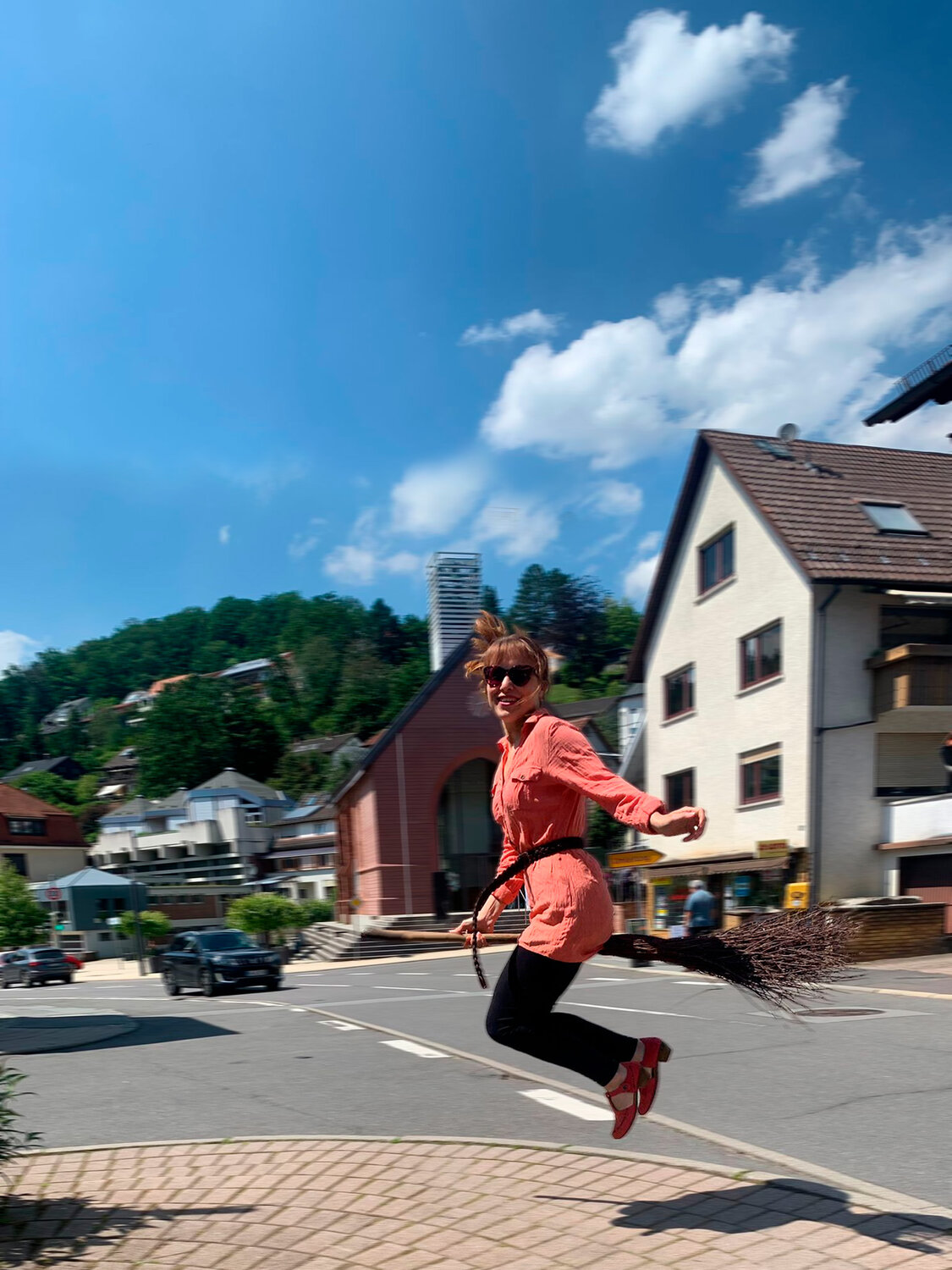 A cheerful young woman in sunglasses jumps high mimicking flying on a traditional wooden broomstick near green trees and houses under a blue sky