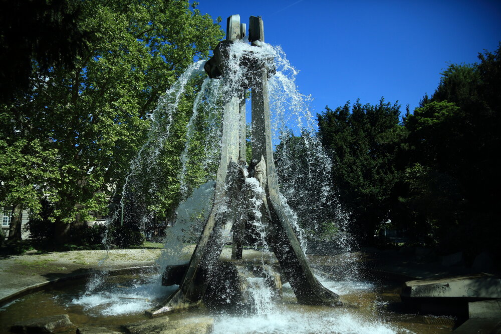 A beautiful detailed wide angle landscape photograph showing a large modern concrete fountain splashing clean water inside a sunny green park sycamore platanus trees area