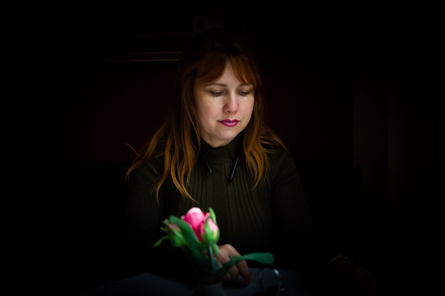 A beautiful detailed close up moody low key portrait photograph showing a thoughtful adult woman sitting inside a dark restaurant next to a pink rose rosa flower during a sunny day
