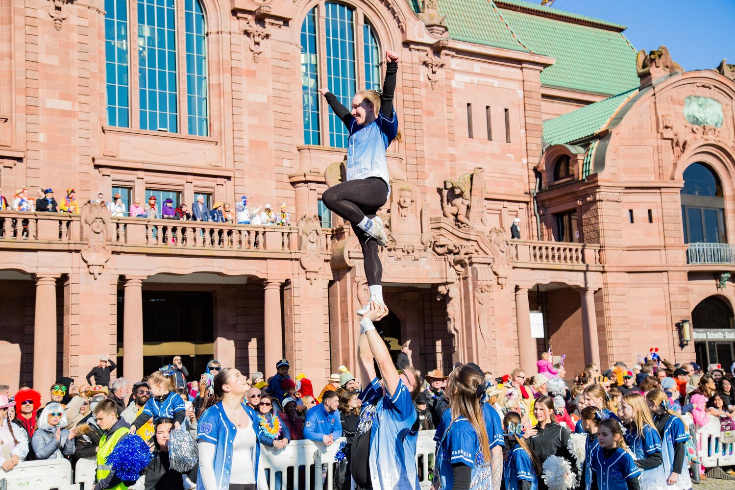 A joyful detailed wide angle action photograph showing cheerleaders performing stunts with many human spectators in front of a historic building during a sunny spring day