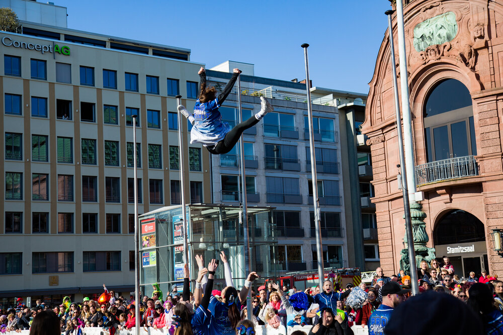 A joyful detailed wide angle action photograph showing cheerleaders performing high aerial acrobatic stunts with many human spectators in front of city buildings during a sunny spring day