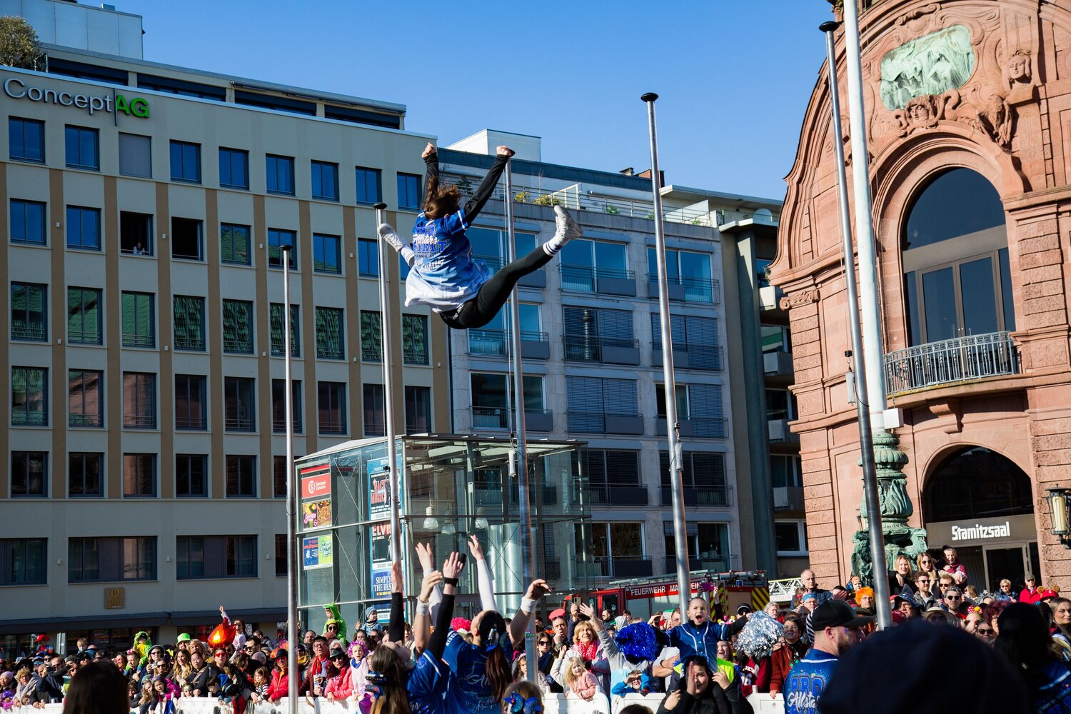 A joyful detailed wide angle action photograph showing cheerleaders performing high aerial acrobatic stunts with many human spectators in front of city buildings during a sunny spring day