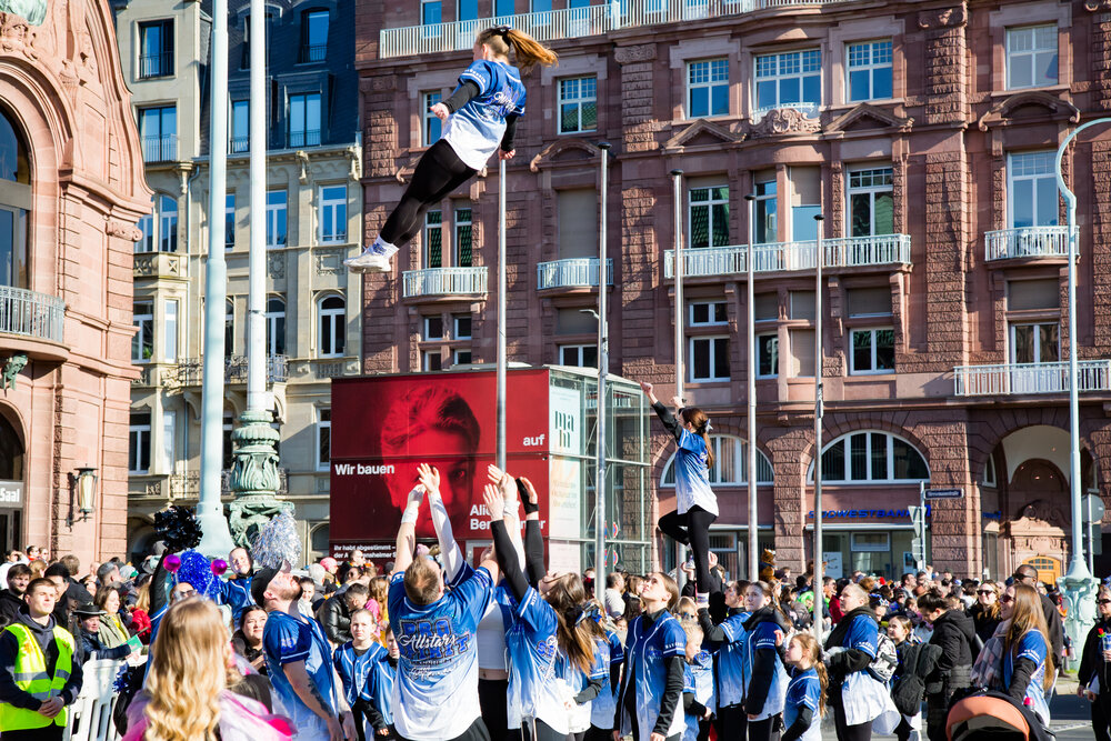 A joyful detailed wide angle action photograph showing young cheerleaders performing high aerial tossed stunts with many human spectators in front of historic buildings during a sunny spring day