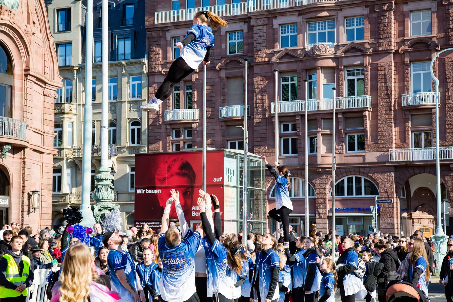A joyful detailed wide angle action photograph showing young cheerleaders performing high aerial tossed stunts with many human spectators in front of historic buildings during a sunny spring day