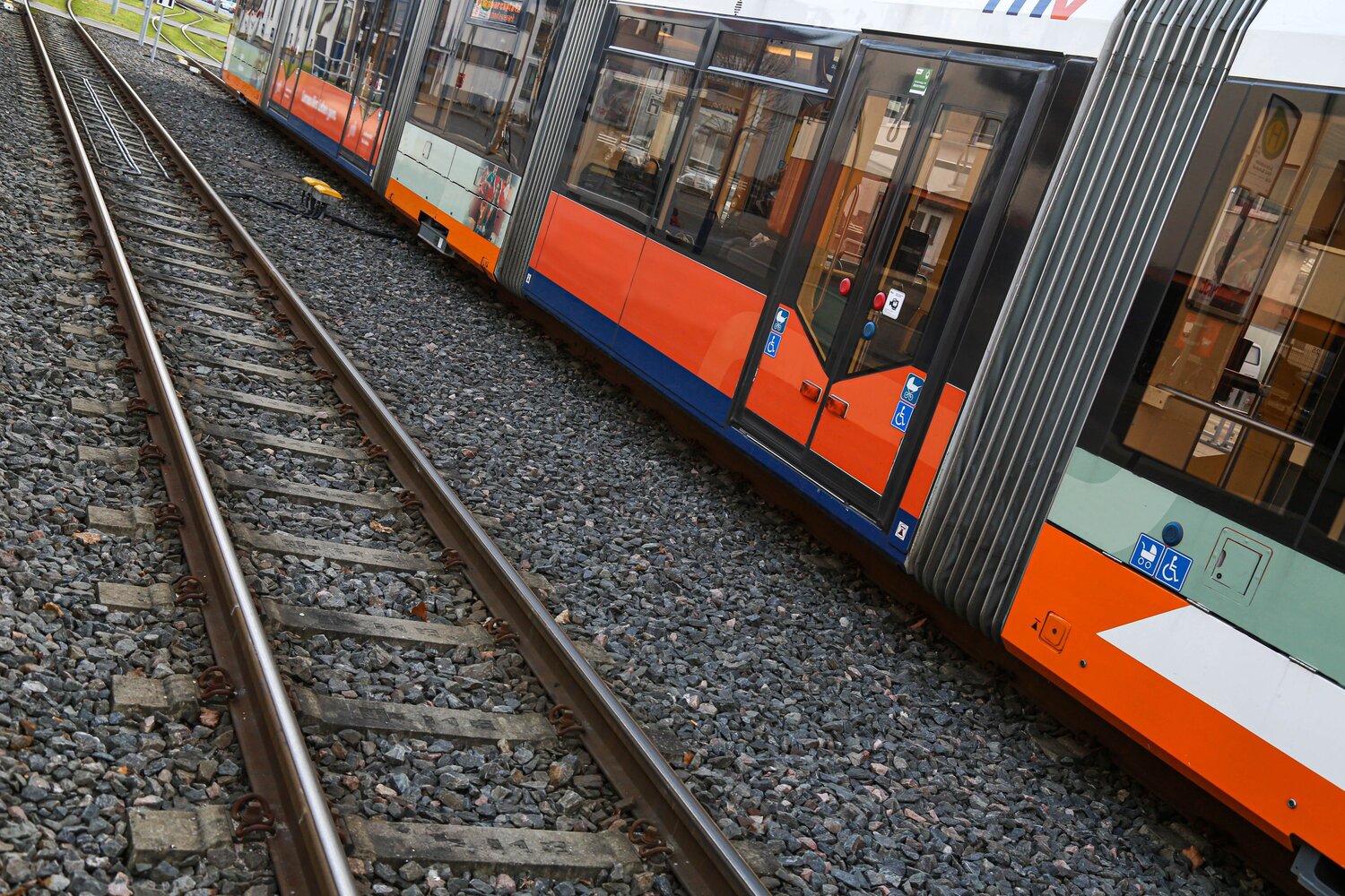A modern orange commuter tram train moving along steel railway tracks with crushed stone ballast background near small roadside grass lawn during a sunny day