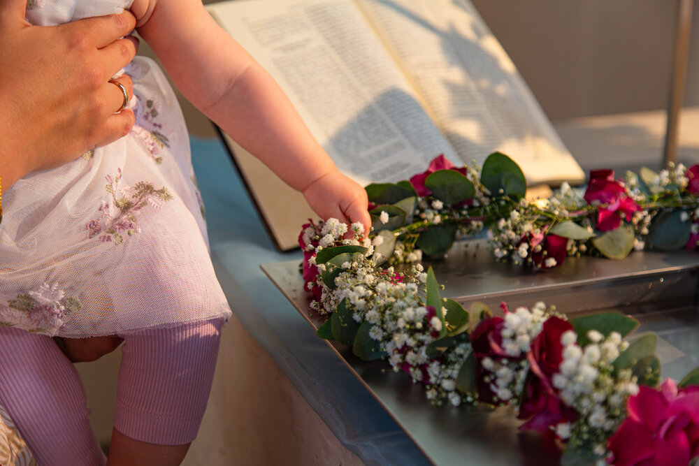 A detailed close up lifestyle photograph showing a little baby hand touching a delicate gypsophila paniculata flower wreath near an open book during a sunny day