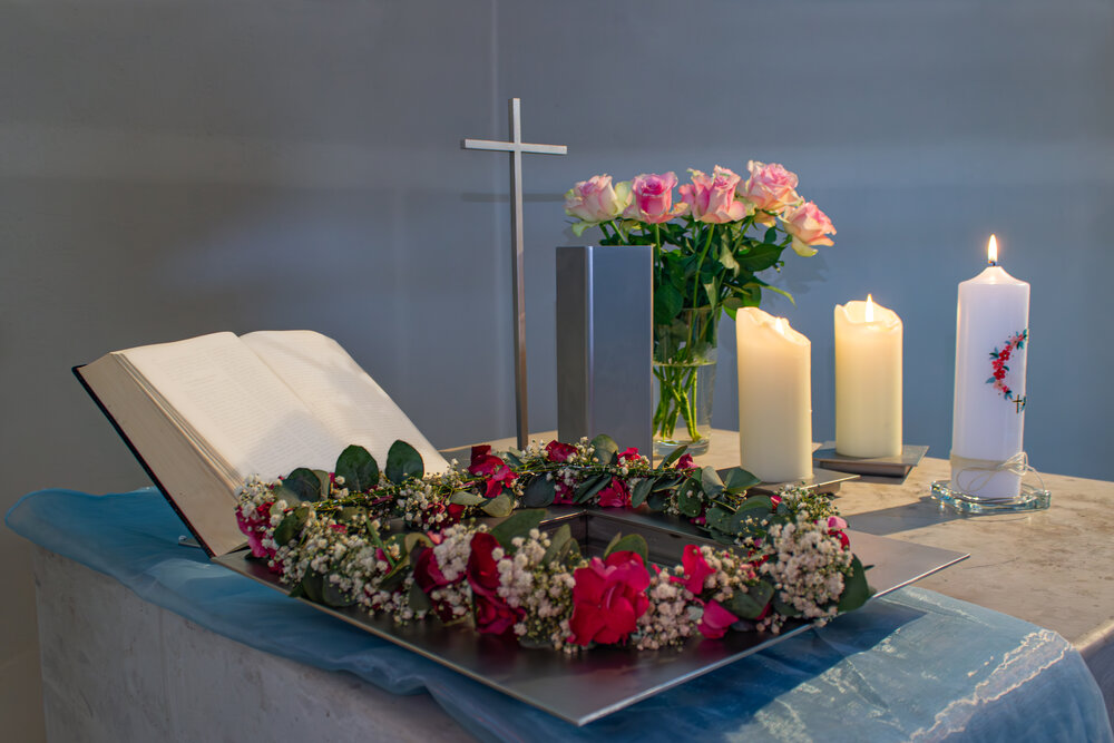 A detailed horizontal interior lifestyle photograph showing a christian church baptismal font holy water vessel with gypsophila wreath open bible cross candles and pink rosa hybrid roses during a sunn