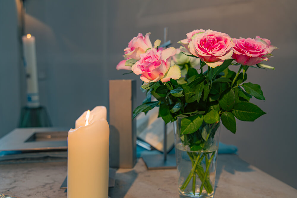 A detailed close up interior lifestyle photograph showing a bouquet of pink rosa hybrid roses in a glass vase with burning white candles inside a church during a sunny day