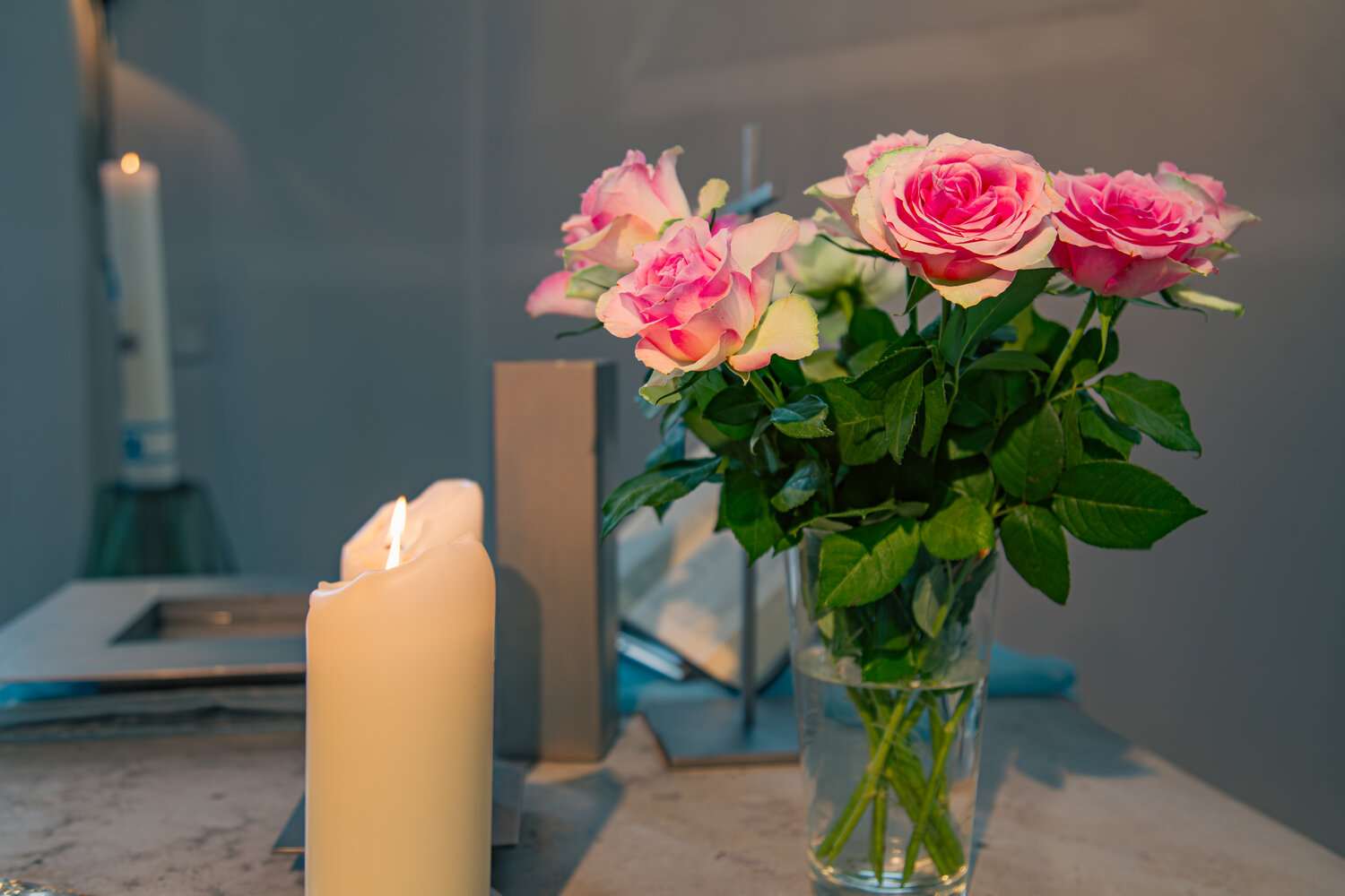 A detailed close up interior lifestyle photograph showing a bouquet of pink rosa hybrid roses in a glass vase with burning white candles inside a church during a sunny day