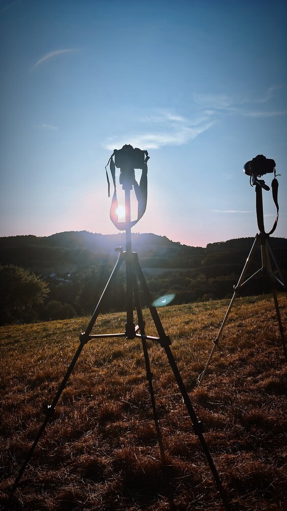 A detailed vertical photograph showing two cameras on tripods shooting sunset landscape silhouette over green hills field