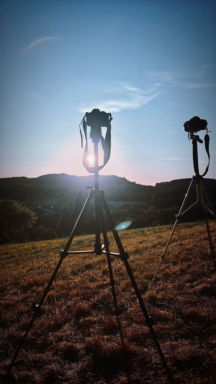 A detailed vertical photograph showing two cameras on tripods shooting sunset landscape silhouette over green hills field