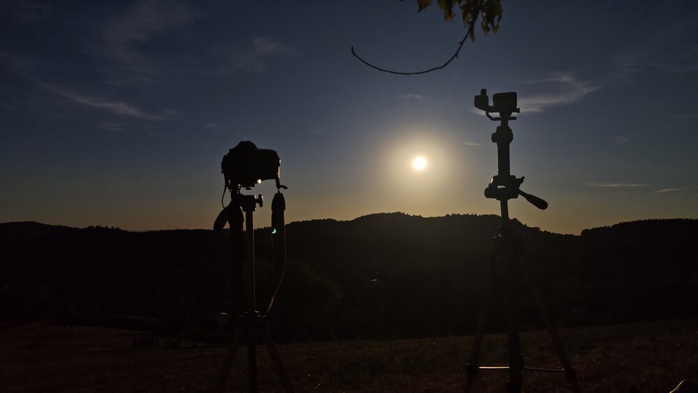 A detailed wide photograph showing camera and action camera on tripods filming dark golden sunset silhouette over mountain hills
