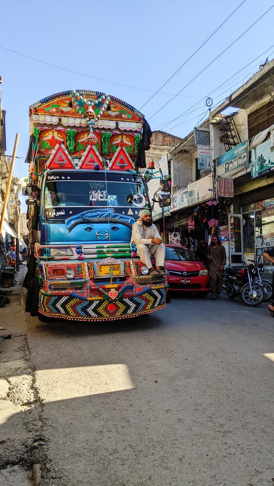 Title: Truck Art Beneath the Hindu Kush  Description: A vibrant piece of Pakistani truck art stands in the heart of Chitral Bazaar, framed by the rugged peaks of the Hindu Kush. The colorful artwork reflects Pakistan’s distinctive folk culture against the