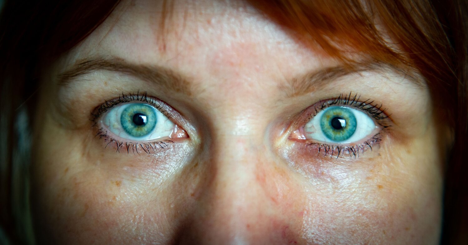 A detailed wide macro portrait showing expressive wide open blue green eyes of a red haired adult woman looking directly at camera