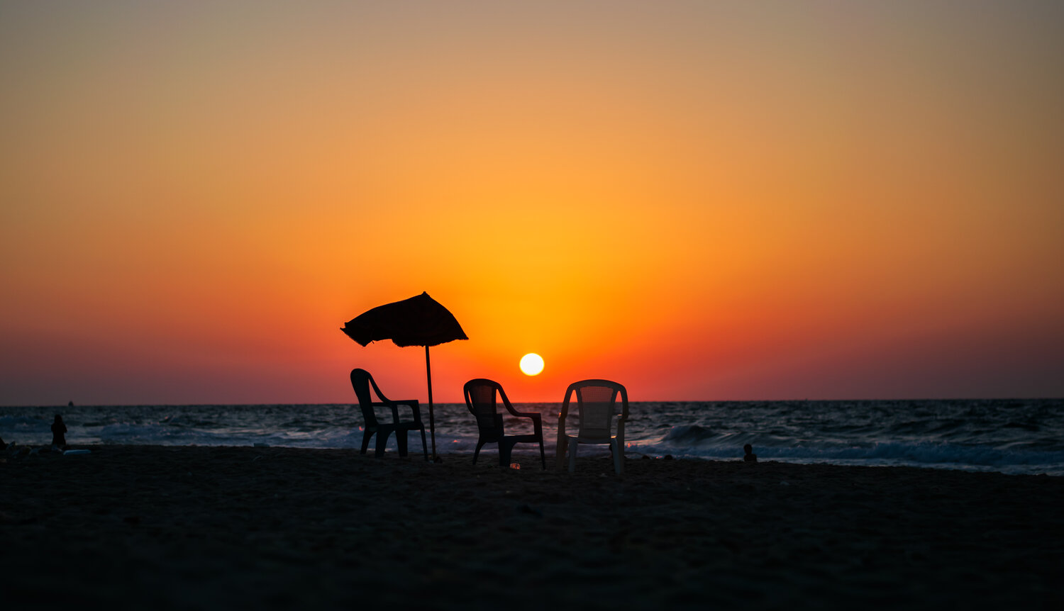 At the end of the day, the beach falls silent beneath a burning sky. Empty chairs and a worn umbrella stand between the fading light and the restless sea, creating a quiet moment where human presence meets the vastness of nature.