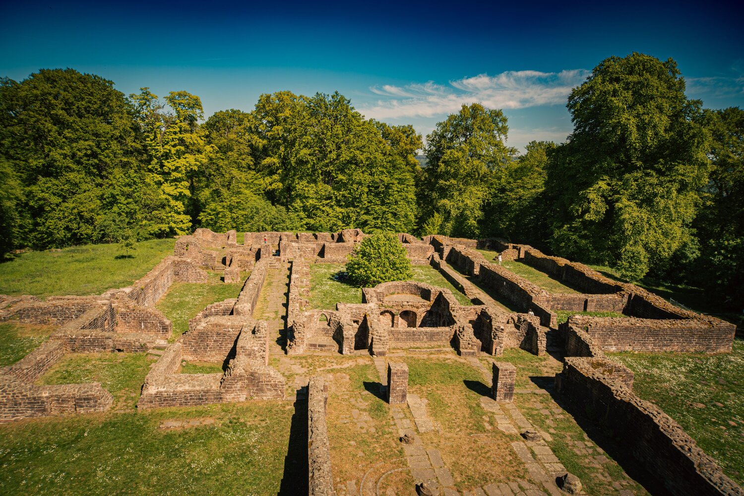 A detailed wide landscape photo showing historic medieval stone ruins of St Michael Monastery surrounded by green forest trees under sunny blue sky