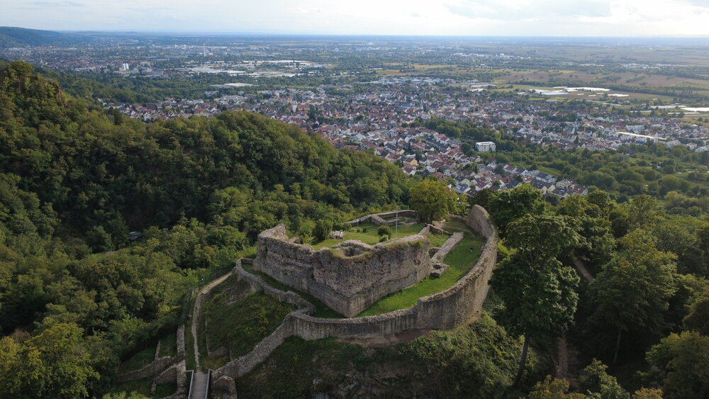 A detailed aerial drone landscape photo showing ancient stone ruins of Strahlenburg Castle on forested mountain ridge overlooking European town valley