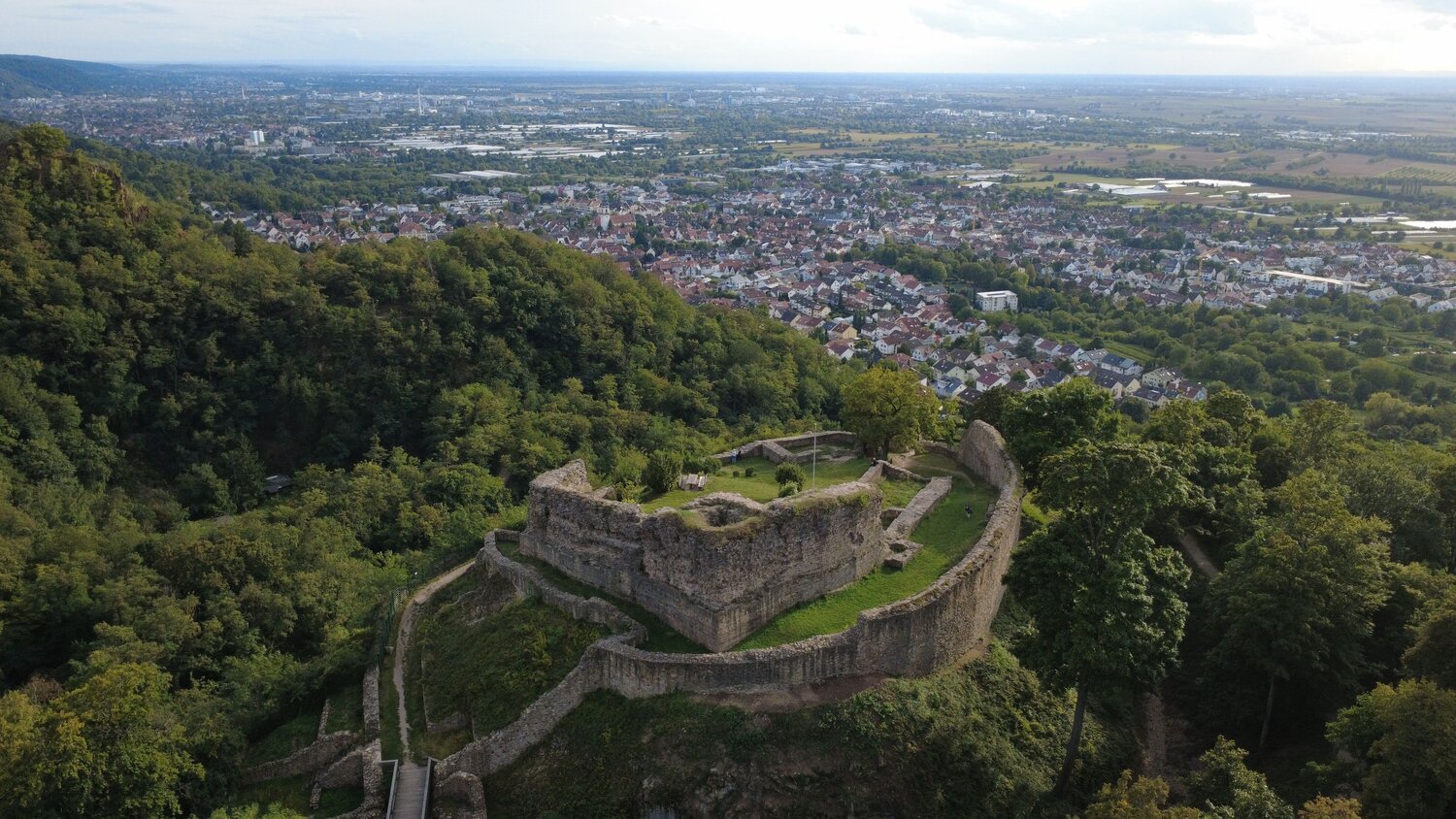 A detailed aerial drone landscape photo showing ancient stone ruins of Strahlenburg Castle on forested mountain ridge overlooking European town valley