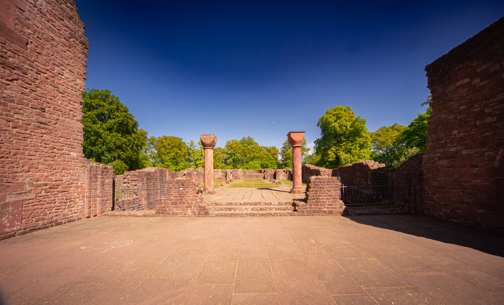 A detailed wide interior landscape photo showing ancient stone ruins of St Michael Monastery with red columns and sandstone walls under blue summer sky