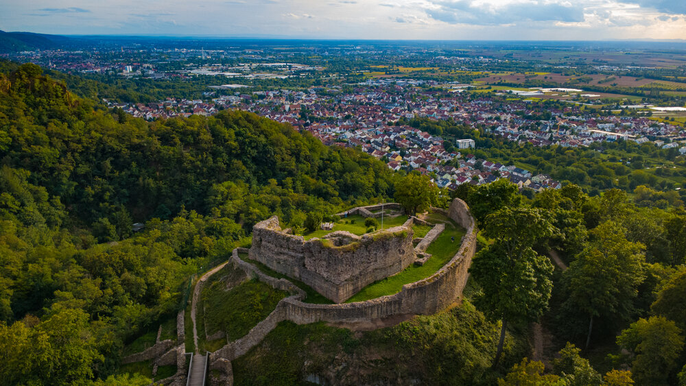 A detailed aerial drone landscape photo showing ancient stone ruins of Strahlenburg Castle on forested mountain ridge overlooking European town valley