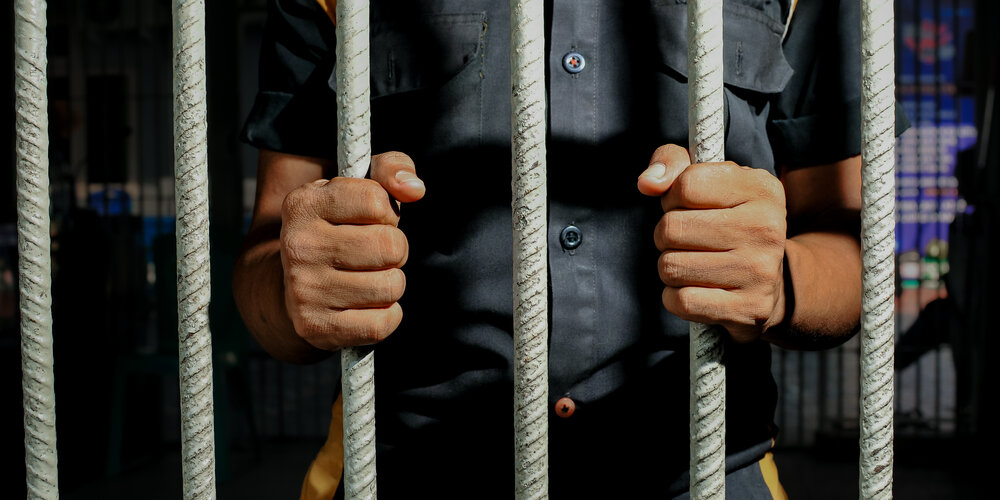 Close up view of hands behind bars in a prison cell, symbolizing incarceration and the criminal justice system.