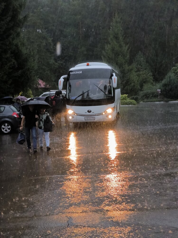 Сцена на улице во время сильного проливного дождя, главным объектом которой является большой белый туристический автобус